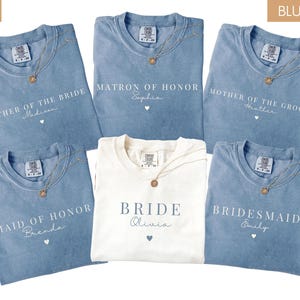 K&ouml;nnte beinhalten: Eine Auswahl an Jeansblauen und elfenbeinfarbenen T-Shirts mit goldenen Halsketten. Die Shirts sind mit Hochzeitsbezeichnungen wie "Mother of the Bride" und "Bridesmaid" sowie Namen bedruckt. Das elfenbeinfarbene Shirt tr&auml;gt die Aufschrift "Bride".