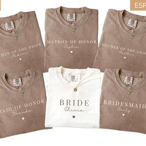 K&ouml;nnte beinhalten: Eine Auswahl an elfenbein- und espressofarbenen T-Shirts mit Aufdrucken f&uuml;r Hochzeitsg&auml;ste. Aufschriften sind "Mother of the Bride", "Matron of Honor", "Mother of the Groom", "Maid of Honor", "Bride" und "Bridesmaid". Jedes Shirt hat eine Halskette.