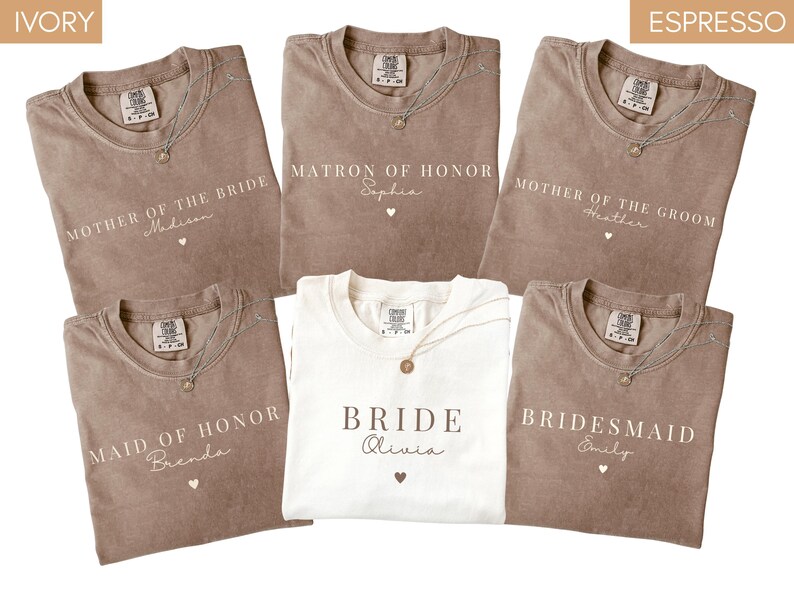 K&ouml;nnte beinhalten: Eine Auswahl an elfenbein- und espressofarbenen T-Shirts mit Aufdrucken f&uuml;r Hochzeitsg&auml;ste. Aufschriften sind "Mother of the Bride", "Matron of Honor", "Mother of the Groom", "Maid of Honor", "Bride" und "Bridesmaid". Jedes Shirt hat eine Halskette.
