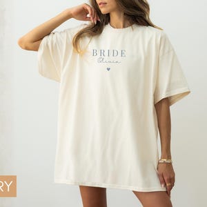 K&ouml;nnte beinhalten: Elfenbeinfarbenes &uuml;bergro&szlig;es T-Shirt mit dem Wort "BRIDE" und einem Namen in zarter Schrift, sowie einer kleinen Herzgrafik. Das Shirt hat kurze &Auml;rmel und eine lockere Passform, ideal f&uuml;r einen l&auml;ssigen Look.