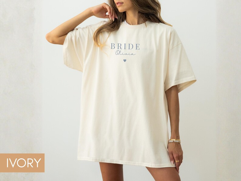 K&ouml;nnte beinhalten: Elfenbeinfarbenes &uuml;bergro&szlig;es T-Shirt mit dem Wort "BRIDE" und einem Namen in zarter Schrift, sowie einer kleinen Herzgrafik. Das Shirt hat kurze &Auml;rmel und eine lockere Passform, ideal f&uuml;r einen l&auml;ssigen Look.