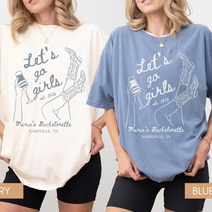 May include: Two t-shirts, one ivory and one blue jean, with the text "Let's go girls" and "Maria's Bachelorette Nashville, TN" printed on them. The design includes a champagne glass and cowboy boots. The shirts are a relaxed fit.