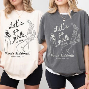 May include: Ivory and pepper-colored t-shirts with a graphic design. The design includes the text "Let's go girls est. 2026" and "Maria's Bachelorette Nashville, TN" with illustrations of a cocktail and cowboy boots.
