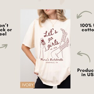 May include: Ivory-colored t-shirt with the text "Let's go girls est. 2024" and a graphic of a woman holding a glass of wine and cowboy boots. The shirt is made of 100% US cotton and is produced in the USA. The text "Maria's Bachelorette Nashville, TN" is also printed on the shirt.