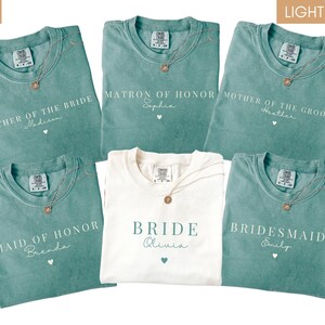 K&ouml;nnte beinhalten: Eine Auswahl an teal- und elfenbeinfarbenen T-Shirts mit goldenen Halsketten. Die teal-farbenen Shirts tragen die Aufschriften "Mother of the Bride", "Matron of Honor", "Mother of the Groom", "Maid of Honor" und "Bridesmaid". Das elfenbeinfarbene Shirt tr&auml;gt die Aufschrift "Bride".