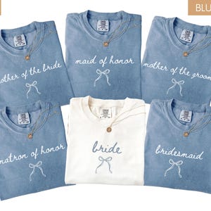 May include: A selection of folded t-shirts in blue jean and ivory. The blue shirts have text that reads "mother of the bride", "maid of honour", "mother of the groom", "matron of honour", and "bridesmaid". The ivory shirt says "bride". Each shirt has a necklace.