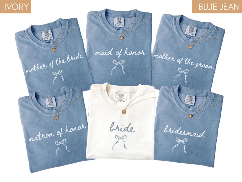 May include: A selection of folded t-shirts in blue jean and ivory. The blue shirts have text that reads "mother of the bride", "maid of honour", "mother of the groom", "matron of honour", and "bridesmaid". The ivory shirt says "bride". Each shirt has a necklace.