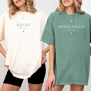 K&ouml;nnte beinhalten: Zwei Frauen tragen &uuml;bergro&szlig;e T-Shirts. Eines ist elfenbeinfarben mit dem Aufdruck "Bride Olivia", das andere hellgr&uuml;n mit dem Aufdruck "Bridesmaid Emily". Beide Shirts haben eine kleine Herzgrafik.