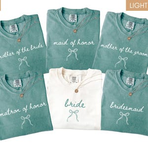 May include: A selection of light green and ivory t-shirts with wedding-related text. The shirts are printed with "mother of the bride," "maid of honour," "mother of the groom," "matron of honour," "bride," and "bridesmaid." Each shirt has a bow graphic.