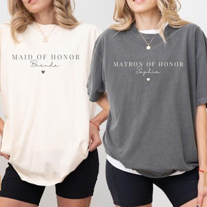K&ouml;nnte beinhalten: Zwei &uuml;bergro&szlig;e T-Shirts, eines in Elfenbein und eines in Pfeffer, mit Text. Das elfenbeinfarbene Shirt tr&auml;gt die Aufschrift "MAID OF HONOR Brenda" mit einem Herz. Das pfefferfarbene Shirt tr&auml;gt die Aufschrift "MATRON OF HONOR Sophia" mit einem Herz. Beide Shirts sind kurz&auml;rmelig.