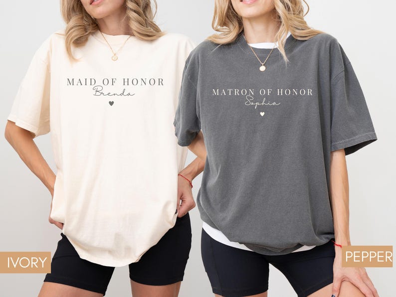 K&ouml;nnte beinhalten: Zwei &uuml;bergro&szlig;e T-Shirts, eines in Elfenbein und eines in Pfeffer, mit Text. Das elfenbeinfarbene Shirt tr&auml;gt die Aufschrift "MAID OF HONOR Brenda" mit einem Herz. Das pfefferfarbene Shirt tr&auml;gt die Aufschrift "MATRON OF HONOR Sophia" mit einem Herz. Beide Shirts sind kurz&auml;rmelig.
