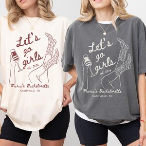 May include: Two oversized t-shirts, one ivory and one pepper, featuring the text "Let's go girls est. 2026" with a graphic of a wine glass and cowboy boots. The text "Maria's Bachelorette Nashville, TN" is also printed on the shirts.
