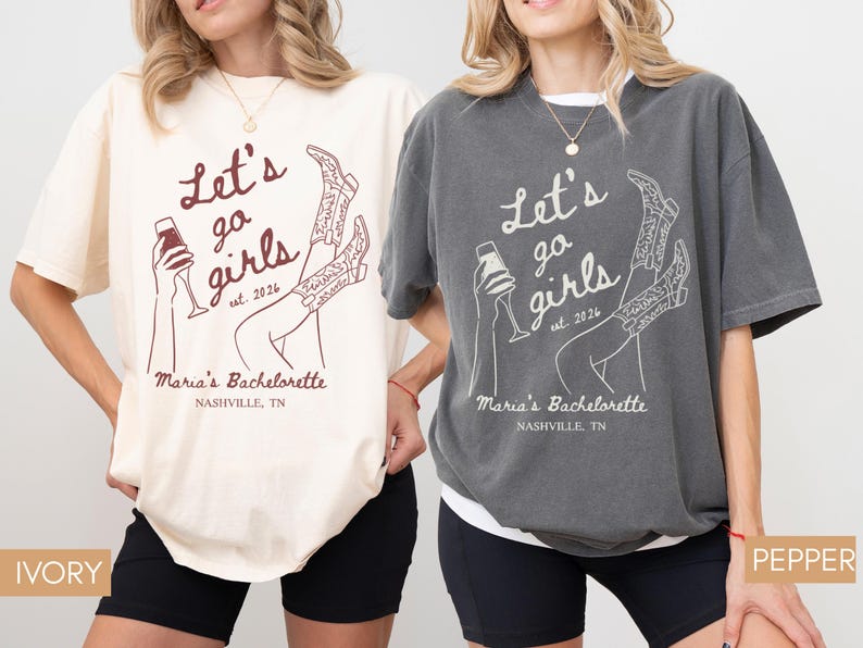 May include: Two oversized t-shirts, one ivory and one pepper, featuring the text "Let's go girls est. 2026" with a graphic of a wine glass and cowboy boots. The text "Maria's Bachelorette Nashville, TN" is also printed on the shirts.