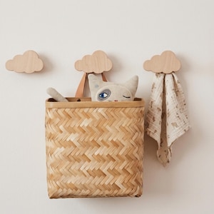 Könnte beinhalten: Drei helle Holzwandhaken in Wolkenform. Ein rechteckiger Korb aus geflochtenem Bambus hängt am mittleren Haken und enthält ein kleines Plüsch-Katzenspielzeug. Ein beiges, gemustertes Stofftuch hängt am rechten Holzhaken in Wolkenform.