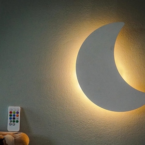 Könnte beinhalten: Eine halbmondförmige Wandleuchte mit warmem, gelbem Licht. Die Lampe ist an einer strukturierten, hellgrauen Wand befestigt. Eine Fernbedienung mit farbigen Tasten ist darunter zu sehen, was auf einstellbare Beleuchtungsoptionen hindeutet. Thema: Heimdekoration.