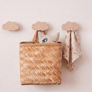Könnte beinhalten: Drei hellfarbene Wandhaken aus Holz in Wolkenform. Ein geflochtener Bambuskorb mit einem Lederriemen hängt am mittleren Haken. Ein Plüsch-Katzenspielzeug mit blauen Augen und einem Augenzwinkern befindet sich im Korb, ein gemustertes Tuch hängt rechts.
