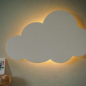 Pode incluir: Uma luz de parede branca em forma de nuvem com um brilho quente atrás. Um controle remoto com várias opções de cores está localizado abaixo. A luz é montada em uma parede verde claro.