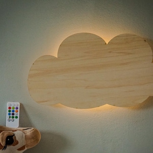 Op de afbeelding: Houten wolkvormige wandlamp met warme LED-verlichting. De lamp is gemonteerd op een lichtgroene muur. Een afstandsbediening met meerdere kleuropties is zichtbaar. Een knuffelgiraf en luiaard staan op de voorgrond.