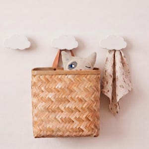Puede incluir: Una cesta de bambú tejida con patrón de espiga cuelga de un gancho de pared blanco en forma de nube. Dentro hay un juguete de peluche de gato con expresión de guiño. Un paño de tela beige con estampado cuelga de un segundo gancho de nube.