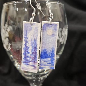 May include: A pair of watercolor earrings featuring a wintery landscape with blue and purple hues. The earrings are rectangular and have silver hooks.