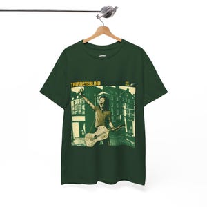 Può includere: T-shirt verde scuro con una grafica di una persona che suona la chitarra davanti a un edificio. Il testo "THIRDEYEBLIND" è stampato in giallo nella parte superiore.