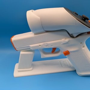 May include: A white 3D-printed VR gun controller with orange accents and a black controller. The controller is mounted on a white stand against a blue background. The design includes a trigger and a grip.