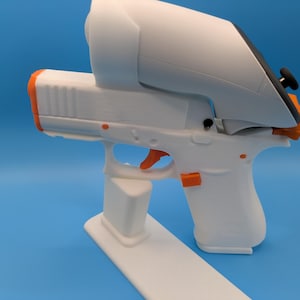 May include: A white and orange VR gun controller with a white stand. The controller has an orange trigger and accents. The VR headset attachment is white and black. The controller is on a blue background.