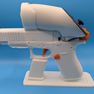 May include: A white and orange toy gun with a VR headset attachment. The toy gun is resting on a white stand. The trigger and other accents are orange. The background is a solid blue.