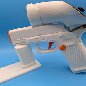 May include: A white and orange toy gun with a white stand and a black and white device attached to the top. The trigger and other details are orange. The background is a solid blue.