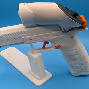 May include: A white and orange 3D-printed gun-shaped controller with a black controller on top. The controller is resting on a white stand with the word "GLOCK" printed on it. The background is a solid blue.