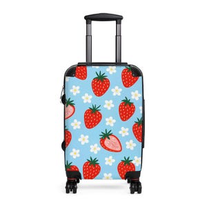 Puede incluir: Una maleta con ruedas con un vibrante estampado de fresas. El diseño presenta fresas rojas con tallos verdes y flores blancas sobre un fondo azul claro. La maleta tiene un asa y ruedas negras.