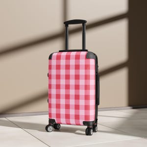 Puede incluir: Una maleta rígida con ruedas que presenta un patrón de cuadros vichy en tonos rosa y rojo. El equipaje incluye un asa retráctil negra, protectores de esquina negros y cuatro ruedas giratorias negras en la base para facilitar el transporte.