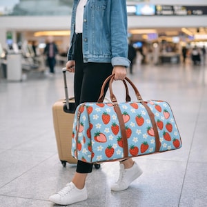 Puede incluir: Una bolsa de viaje azul con un estampado de fresas y flores blancas. La bolsa tiene asas y correas de cuero marrón. Una maleta beige con ruedas está al lado de la bolsa.