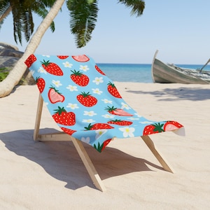 Puede incluir: Una silla de playa de madera con tela azul claro con un estampado de fresas rojas y flores blancas. La silla está en una playa de arena con un barco al fondo. Ideal para relajarse al aire libre.