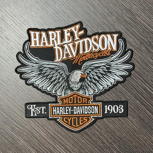 Può includere: Toppa ricamata nera e arancione con un logo di aquila Harley-Davidson. L'aquila ha le ali bianche e sta volando con le ali spiegate. Il testo "Harley-Davidson Motorcycles" è sopra l'aquila e il testo "Motor? Harley-Davidson? Cycles" è sotto l'aquila. Il testo "1903" è sul lato destro della toppa.