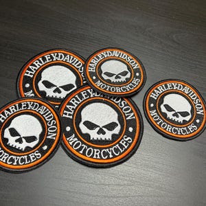 Peut inclure: Plusieurs écussons Harley-Davidson Motorcycles. Chaque écusson est circulaire avec une bordure noire, un anneau intérieur orange et du texte blanc. Le motif central représente un crâne blanc. Les écussons varient en taille.