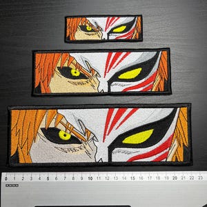 Könnte beinhalten: Drei gestickte Aufnäher mit einem Anime-Design von Augen und einer Maske. Die Aufnäher haben die Farben Orange, Weiß, Schwarz und Gelb. Die Aufnäher sind rechteckig mit schwarzen Rändern. Ein Lineal liegt darunter.
