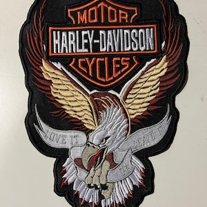 Op de afbeelding: Geborduurde Harley-Davidson patch met een adelaar met uitgespreide vleugels. De patch heeft een zwarte achtergrond met het Harley-Davidson logo in oranje en wit. De adelaar houdt een banner met de tekst "LOVE IT OR LEAVE IT".