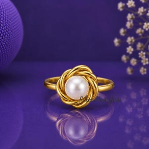 May include: A gold-toned ring featuring a central white pearl surrounded by a decorative, twisted metal knot design. The band is smooth and circular, showcasing a polished finish that reflects light, creating a clear, symmetrical appearance.