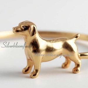 Puede incluir: Un anillo dorado con una detallada figura de perro. El perro está de pie sobre sus cuatro patas, con la cola levantada. La banda del anillo es de diseño simple y delgado. El anillo es probablemente de metal.