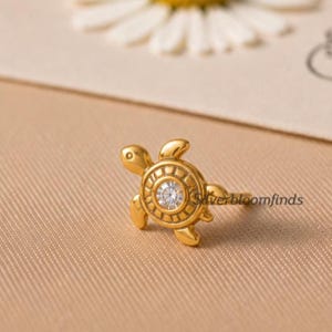 Puede incluir: Un anillo dorado con forma de tortuga con una gema redonda y transparente en el centro del caparazón. El anillo está sobre un fondo neutro. Es una joya.