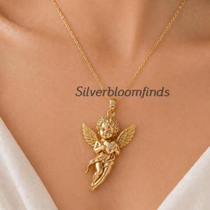 May include: Gold-colored necklace featuring a cherub pendant holding a heart. The cherub has detailed wings and is suspended from a delicate chain. The text "Silverbloomfinds" is visible on the image.
