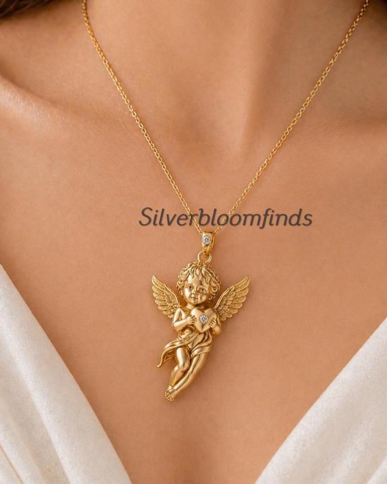 May include: Gold-colored necklace featuring a cherub pendant holding a heart. The cherub has detailed wings and is suspended from a delicate chain. The text "Silverbloomfinds" is visible on the image.