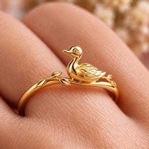 Könnte beinhalten: Ein goldfarbener Ring mit einer kleinen Entenfigur, die auf einem dünnen, astförmigen Ringband sitzt. Die Ente hat detaillierte Flügelstrukturen und einen glatten Kopf, während das Band kleine blattförmige Akzente aufweist.