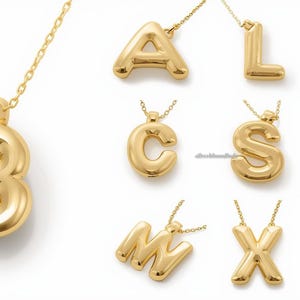 Może przedstawiać: Złote naszyjniki z inicjałami. Obraz przedstawia różnorodne naszyjniki w odcieniach złota, każdy z inną literą alfabetu. Litery są w zaokrąglonej, bąbelkowej czcionce i zawieszone na delikatnych złotych łańcuszkach.