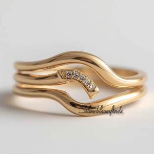 May include: A gold-colored ring set with a unique, flowing design. The ring features a curved band with a section of small, clear gemstones. The design is elegant and modern, suitable for everyday wear or special occasions.