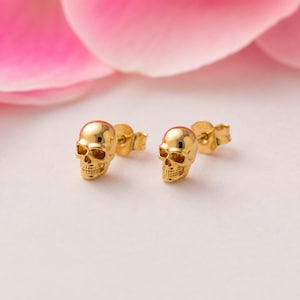 May include: A pair of gold-toned skull stud earrings. Each earring features a detailed, polished skull design with a post back. The back of each earring is clearly stamped with the text 925, indicating the metal composition of the jewelry pieces.