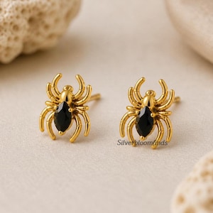 May include: A pair of gold-colored spider stud earrings. Each earring features a spider design with a black gemstone body. The earrings are displayed on a neutral background.