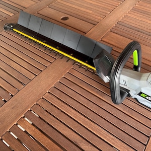 Puede incluir: Un cortasetos inalámbrico con protector de cuchilla negro y cuerpo gris y negro. El cortasetos tiene un mango negro con un acento verde y una guía de cuchilla amarilla. La marca "EGO" es visible en el lateral.