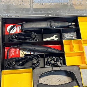 May include: A black and yellow tool kit with a clear lid. The kit contains two black rotary tools, a small white wrench, and various accessories. Yellow compartments hold additional items, including small gold-colored shapes.